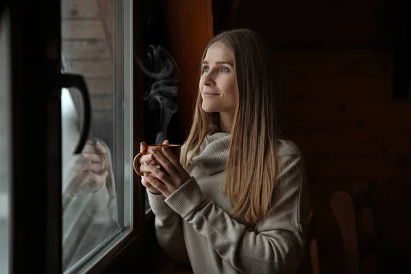 Mujer joven con bebida caliente cerca de la ventana en el día de invierno —  Fotos de Stock
