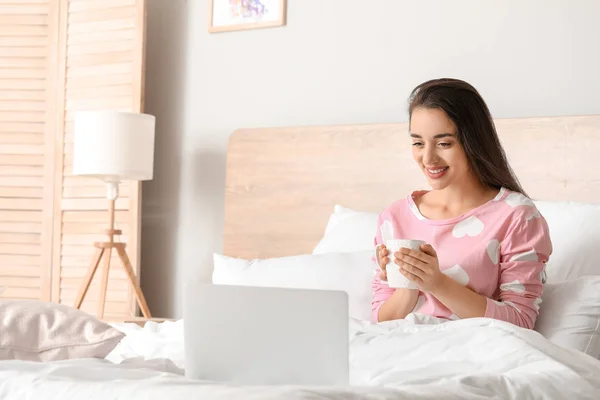 Morning of beautiful young woman with laptop and cup of coffee in bedroom — Stock Photo, Image
