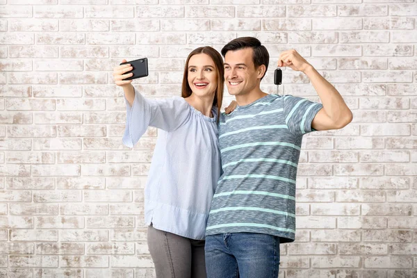 Feliz pareja con la llave de su nuevo coche tomando selfie en el fondo de ladrillo —  Fotos de Stock