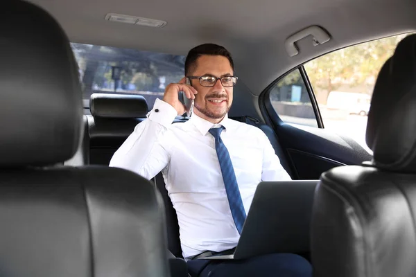 Successful businessman with laptop talking by phone in modern car — Stock Photo, Image
