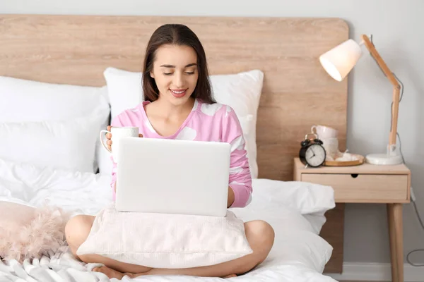 Morning of beautiful young woman with laptop and cup of coffee in bedroom — Stock Photo, Image