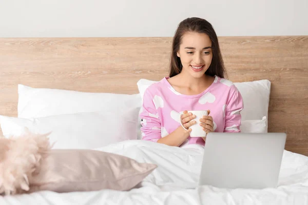 Morning of beautiful young woman with laptop and cup of coffee in bedroom — Stock Photo, Image