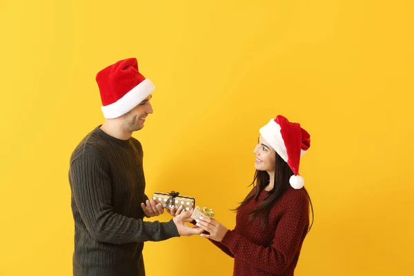 Happy young man and woman giving each other Christmas presents against color background — Stock Photo, Image