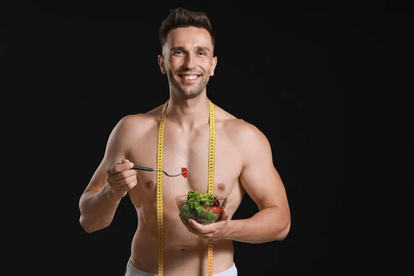 Handsome muscular man with measuring tape and salad on dark background. Weight loss concept — Stock Photo, Image