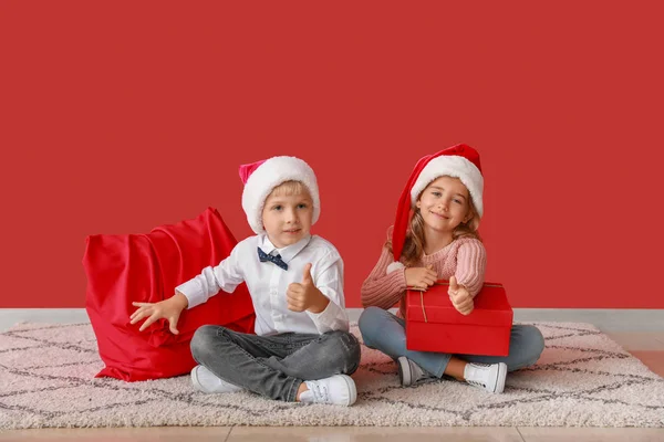 Söta små barn i Santa Claus hattar och med julklappar som visar tummen upp gest nära färg väggen — Stockfoto
