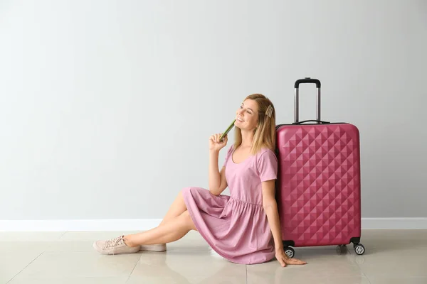 Happy beautiful woman with luggage and passport sitting near light wall — Stock Photo, Image
