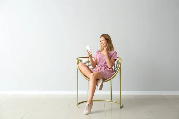 Happy beautiful with mobile phone sitting in armchair near light wall — Stock Photo, Image