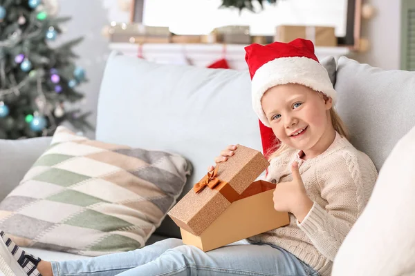 Cute little girl with Christmas gift showing thumb-up at home — Stock Photo, Image