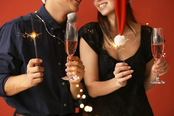 Ευτυχισμένο νεαρό ζευγάρι με χριστουγεννιάτικα βεγγαλικά και ποτήρια σαμπάνιας σε έγχρωμο φόντο — Φωτογραφία Αρχείου