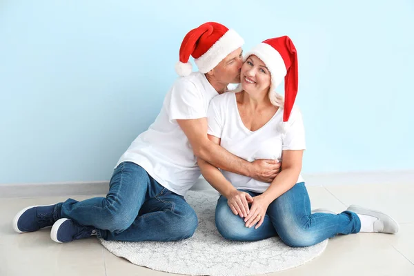 Happy mature couple in Santa Claus hats sitting near light wall — Stock Photo, Image