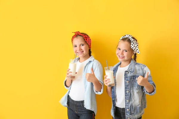 Portrait of cute twin girls with glasses of milk showing thumb-up gesture on color background — Stock Photo, Image