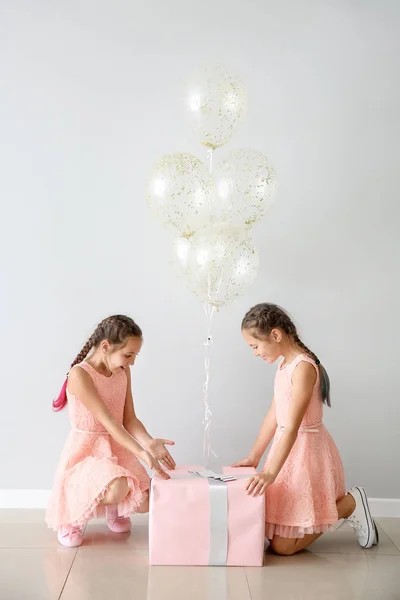 Portrait of cute twin girls with air balloons and big gift near light wall — Stock Photo, Image