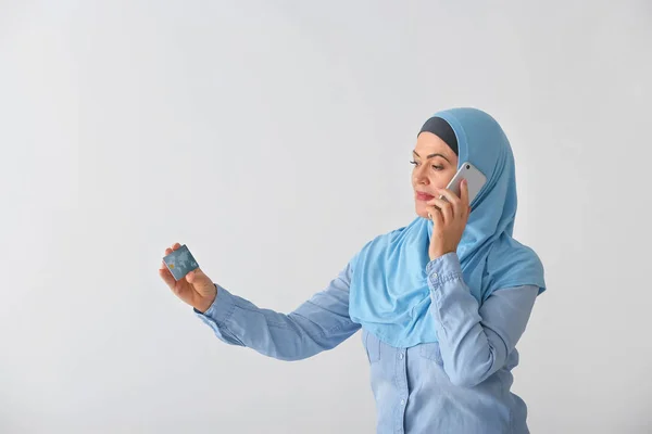 Beautiful mature Muslim woman with credit card talking by mobile phone on light background — Stock Photo, Image