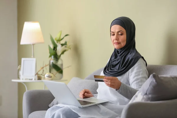 Gyönyörű érett muszlim nő laptoppal és bankkártyával otthon — Stock Fotó