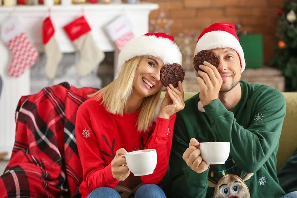 Pareja feliz en los sombreros de Santa beber chocolate caliente y comer galletas en casa — Foto de Stock