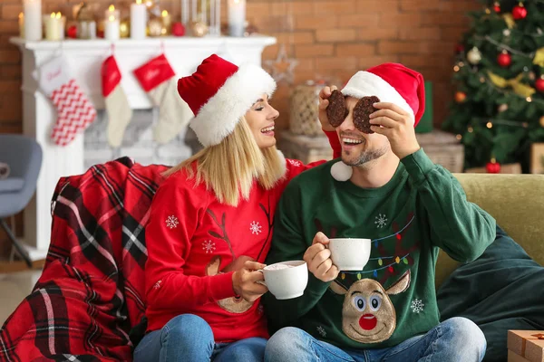 Casal feliz em chapéus de Santa beber chocolate quente e comer biscoitos em casa — Fotografia de Stock