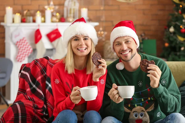 Happy couple in Santa hats drinking hot chocolate and eating cookies at home — Stock Photo, Image