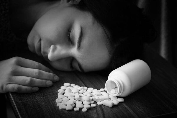 Black and white photo of young unconscious woman with pills on table. Suicide concept
