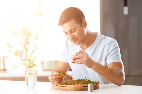 Молодой человек солит вкусную пиццу за столом — стоковое фото