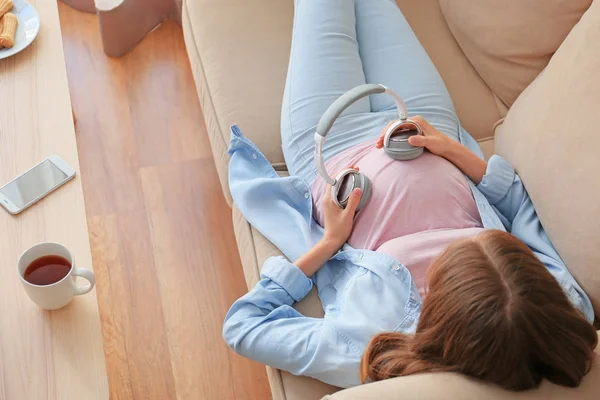 Όμορφη έγκυος γυναίκα βάζοντας τα ακουστικά στην κοιλιά της, ενώ βρίσκεται στον καναπέ — Φωτογραφία Αρχείου
