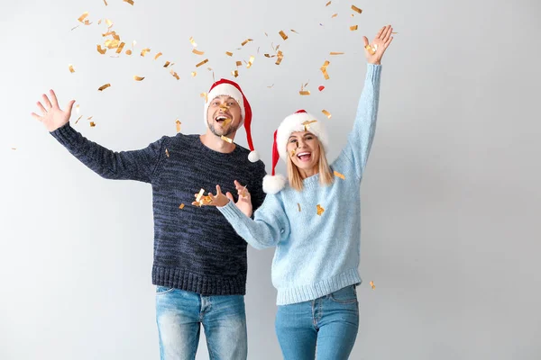Happy couple in Santa hats and falling confetti on light background — Stock Photo, Image