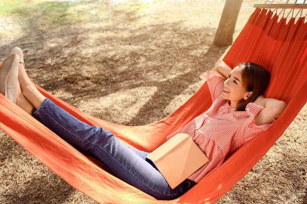 Красива молода жінка з книгою відпочиває в гамаку на відкритому повітрі — стокове фото