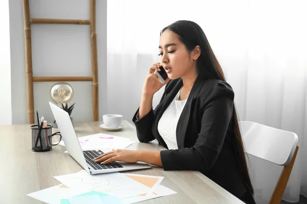 Beautiful Asian businesswoman with laptop talking by mobile phone in office — Stock Photo, Image