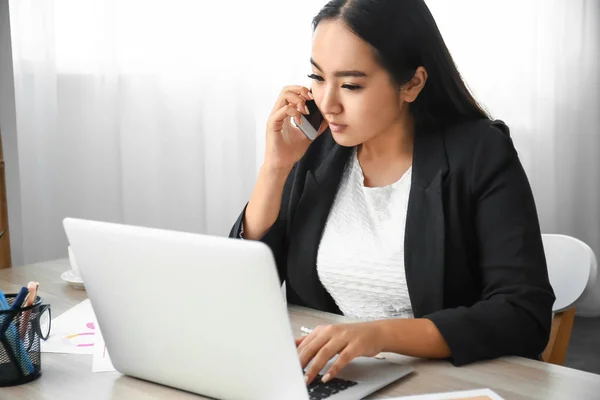 Beautiful Asian businesswoman with laptop talking by mobile phone in office — Stock Photo, Image