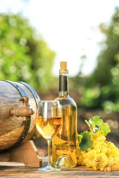 Bicchiere e bottiglia di vino bianco con uva fresca e botte sul tavolo di legno in vigna — Foto Stock