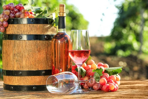 Bicchieri e bottiglia di vino con uva fresca e botte sul tavolo di legno in vigna — Foto Stock
