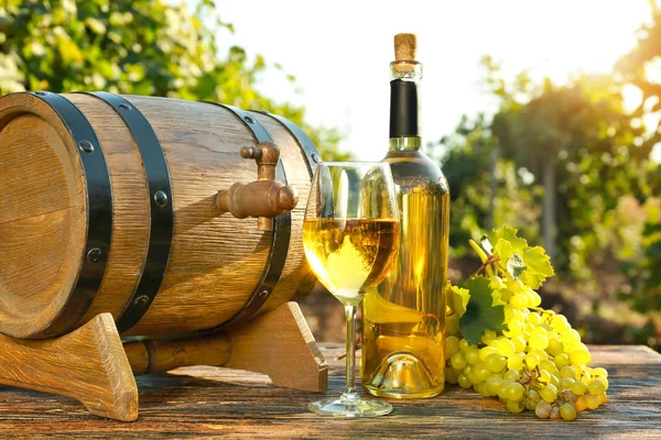 Copa y botella de vino blanco con uvas frescas y barril sobre mesa de madera en viñedo —  Fotos de Stock