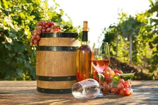 Glazen en fles wijn met verse druiven en vat op houten tafel in wijngaard — Stockfoto