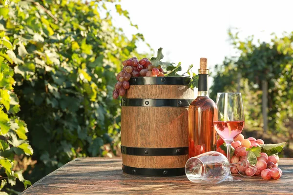 Glazen en fles wijn met verse druiven en vat op houten tafel in wijngaard — Stockfoto