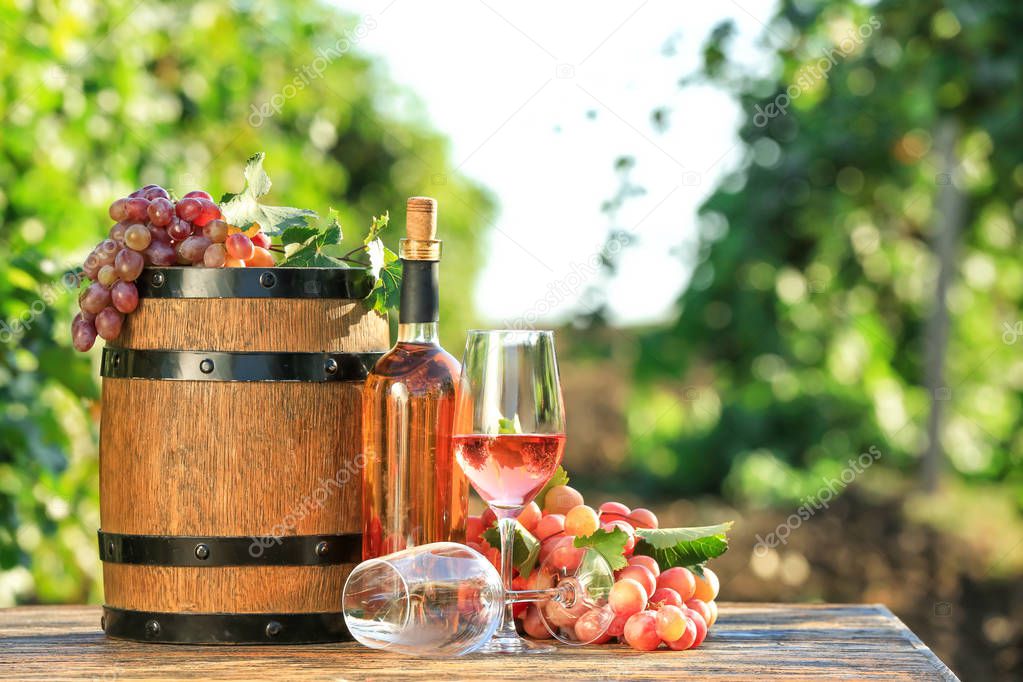 Glasses and bottle of wine with fresh grapes and barrel on wooden table in vineyard