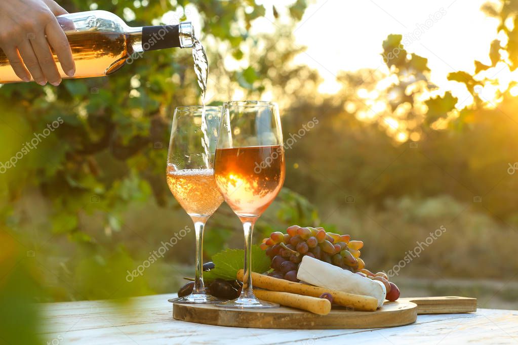 Pouring of tasty wine from bottle into glasses on table with snacks in vineyard