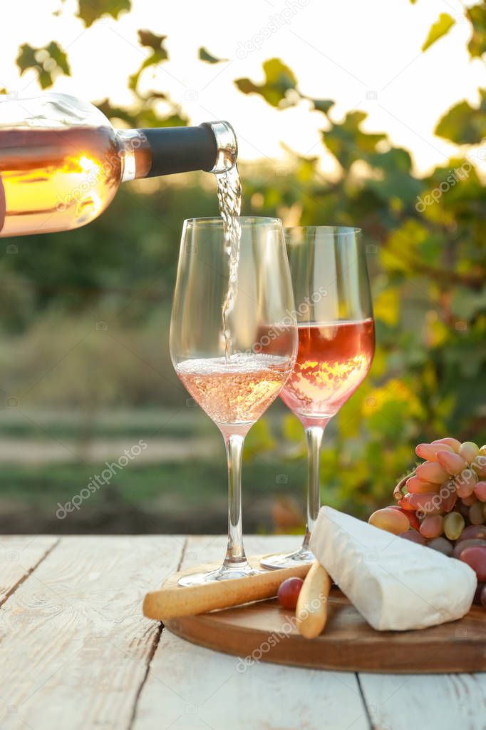 Pouring of tasty wine from bottle into glasses on table with snacks in vineyard
