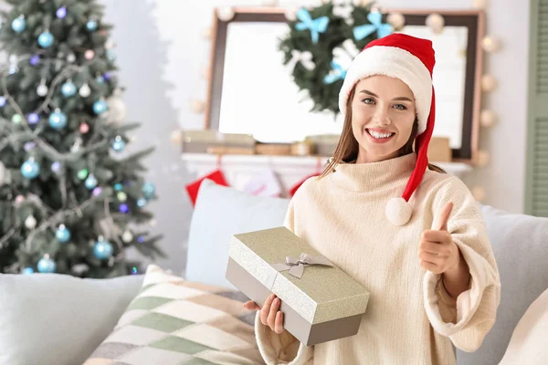 Beautiful young woman with Christmas gift showing thumb-up at home — Stock Photo, Image