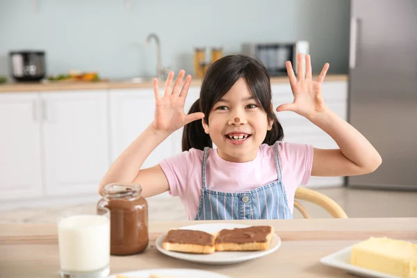 Funny little girl eating tasty toasts with chocolate spreading in kitchen — Stock Photo, Image