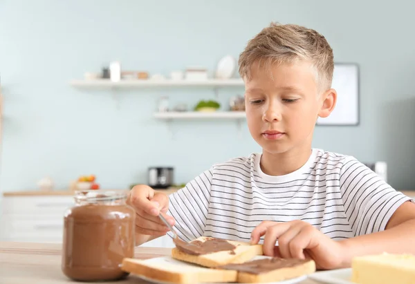 Funny little boy eating tasty toasts with chocolate spreading in kitchen — Stock Photo, Image