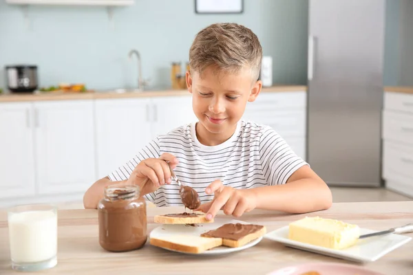 Funny little boy eating tasty toasts with chocolate spreading in kitchen — Stock Photo, Image