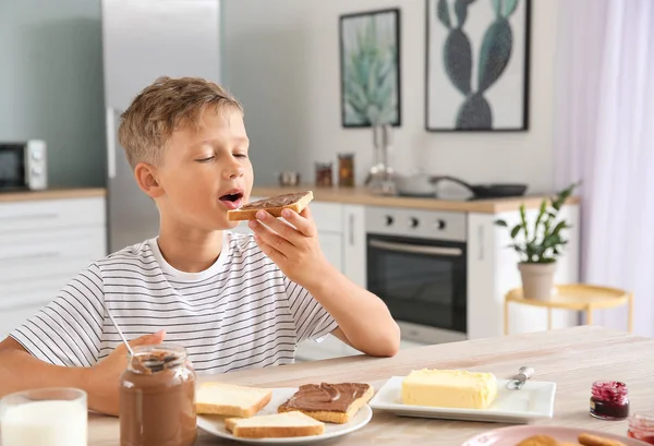 Funny little boy eating tasty toasts with chocolate spreading in kitchen — Stock Photo, Image