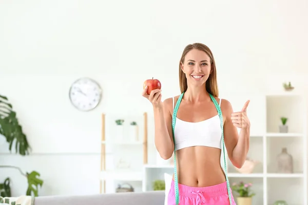 Beautiful young woman with measuring tape and apple showing thumb-up at home. Weight loss concept — Stock Photo, Image