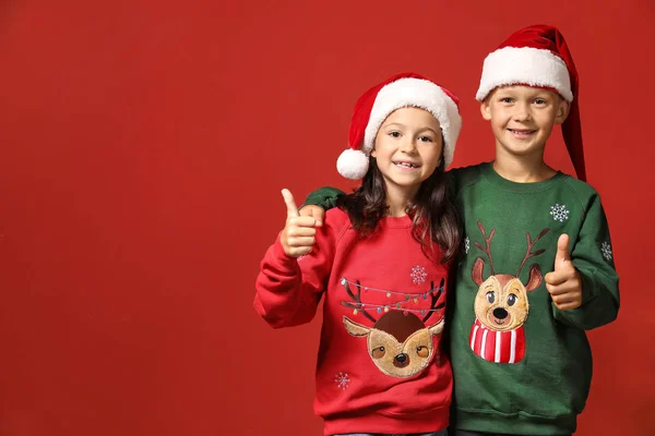 Söta barn i jultröjor och Santa hattar visar tummen upp på färg bakgrund — Stockfoto