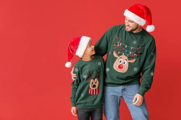Homem feliz com seu filho em camisolas de Natal e chapéus de Papai Noel no fundo de cor — Fotografia de Stock