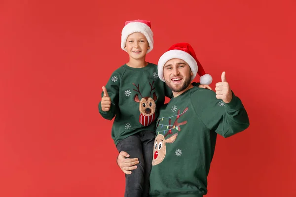 Happy man with his son in Christmas sweaters and Santa hats showing thumb-up on color background — Stock Photo, Image