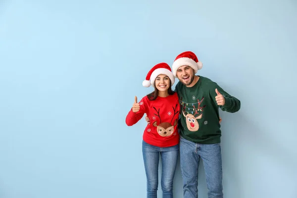 Pareja feliz en suéteres de Navidad y sombreros de Santa mostrando el pulgar hacia arriba en el fondo de color — Foto de Stock