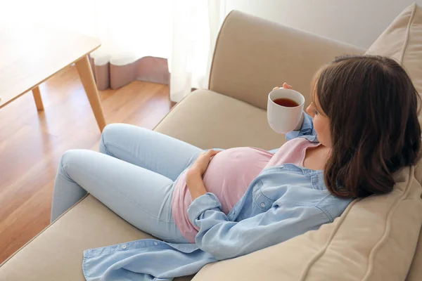 Beautiful pregnant woman drinking tea while sitting on sofa at home — Stock Photo, Image