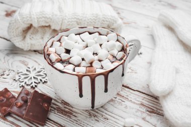 Bir fincan kremalı sıcak çikolata, örgü şapka ve beyaz ahşap arka planda eldivenler.