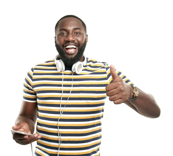 Happy African-American man with mobile phone and headphones showing thumb-up gesture on white background — Stock Photo, Image