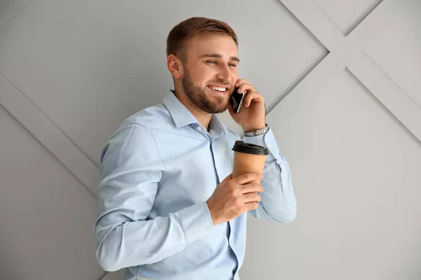 Portrait of handsome businessman with cup of coffee talking by phone on light background — Stock Photo, Image